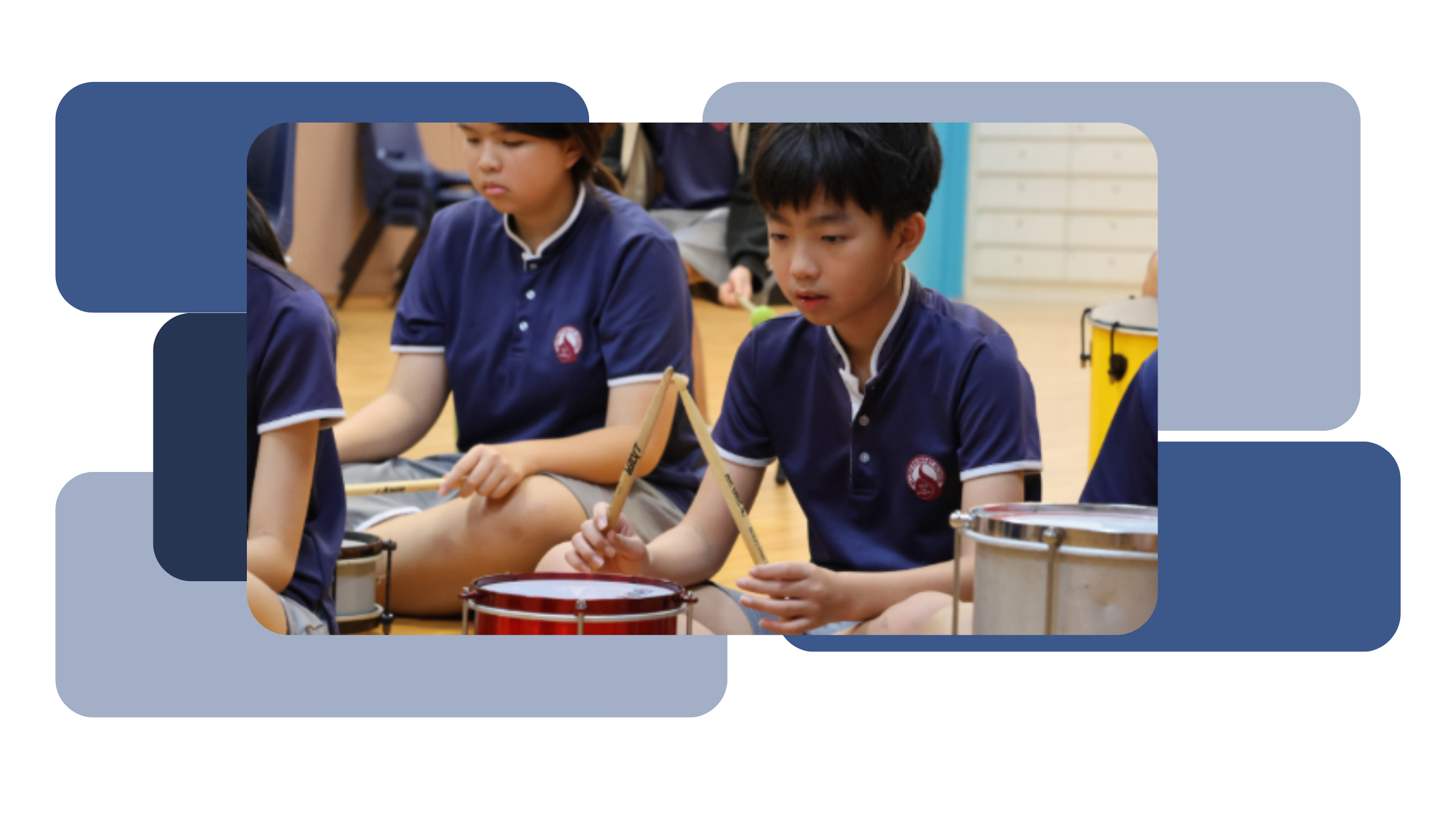 Students in school uniforms participating in a Brazilian Drumming activity, seated on the floor and playing drums with drumsticks, with various drums including a red snare drum and a yellow conga drum visible in the background.